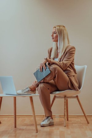 Foto de Una mujer de negocios profesional se sienta en una silla, rodeada de un fondo beige sereno, trabajando diligentemente en su computadora portátil, mostrando dedicación y enfoque en su búsqueda del éxito. - Imagen libre de derechos