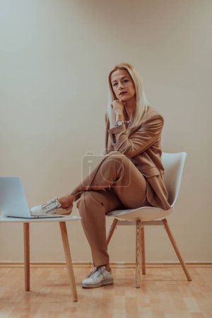 Foto de Una mujer de negocios profesional se sienta en una silla, rodeada de un fondo beige sereno, trabajando diligentemente en su computadora portátil, mostrando dedicación y enfoque en su búsqueda del éxito. - Imagen libre de derechos