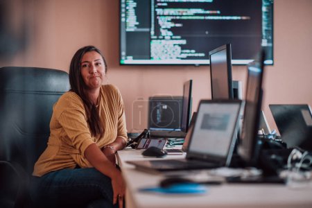 Foto de Una mujer de negocios sentada en una oficina de programadores rodeada de computadoras, mostrando su experiencia y dedicación a la tecnología - Imagen libre de derechos