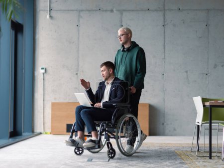 Foto de Un hombre de negocios en silla de ruedas en una oficina de moda usando un portátil mientras está detrás de él es su colega de negocios que le da apoyo. - Imagen libre de derechos