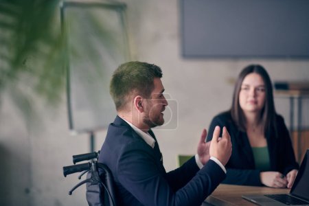 Photo for A wheelchair bound businessman confidently leads a business meeting in a large, modern office, exemplifying inclusive leadership, effective communication, and the power of diversity in driving success - Royalty Free Image
