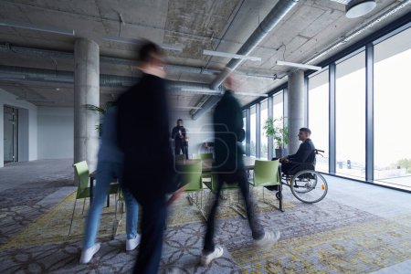 Téléchargez les photos : Un homme d'affaires en fauteuil roulant est assis à une table dans un grand bureau moderne, tandis que ses collègues se rassemblent autour, leurs pas flous, symbolisant l'inclusion, le soutien et l'unité face aux défis - en image libre de droit