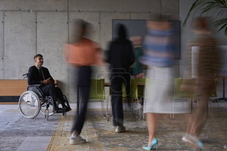 Téléchargez les photos : Un homme d'affaires en fauteuil roulant dans un bureau moderne, entouré de ses collègues qui sont représentés avec des mouvements flous, symbolisant leur soutien et leur solidarité alors qu'ils naviguent dans l'espace de travail - en image libre de droit