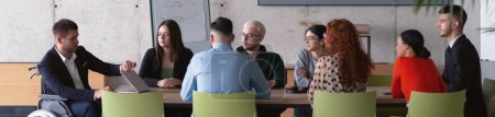 Wide crop photo of a diverse group of business professionals, including an person with a disability, gathered at a modern office for a productive and inclusive meeting