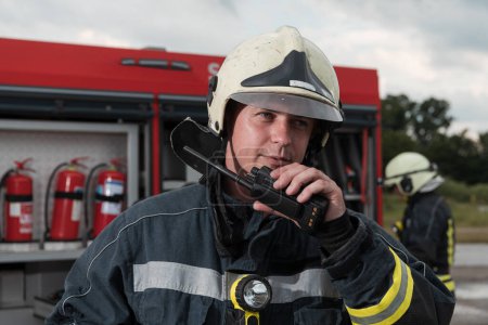 Téléchargez les photos : Pompier utilisant talkie walkie au camion de pompiers d'action de sauvetage et l'équipe de pompiers en arrière-plan. Photo de haute qualité - en image libre de droit