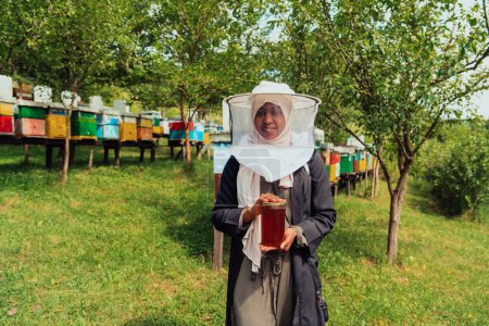Foto de Portrait of a Muslim African American woman in the beekeeping department of a honey farm holding a jar of honey in her hand. - Imagen libre de derechos