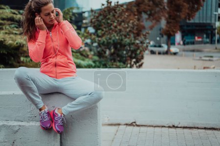 Foto de A woman in a sports outfit is resting in a city environment after a hard morning workout while using noiseless headphones. - Imagen libre de derechos