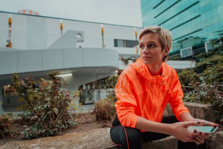 Téléchargez les photos : Une femme en tenue de sport assise dans un environnement urbain et utilisant un smartphone après une dure séance d'entraînement matinale. - en image libre de droit