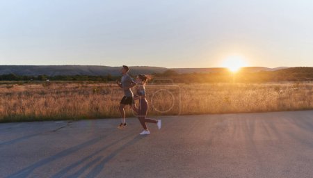 Foto de Una hermosa pareja joven corriendo juntos durante las primeras horas de la mañana, con el fascinante amanecer emitiendo un cálido resplandor, simbolizando su amor y vitalidad compartidos. - Imagen libre de derechos