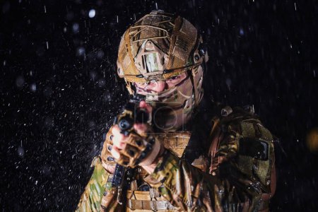 Téléchargez les photos : Soldat de l'armée en uniforme de combat avec un fusil d'assaut, un porte-plaque et un casque de combat en mission dangereuse par une nuit pluvieuse - en image libre de droit
