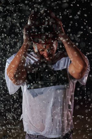 Téléchargez les photos : American Football Field : Solitaire athlète guerrier debout sur un terrain tient son casque et prêt à jouer. Joueur se préparant à courir, attaquer et marquer Touchdown. Nuit pluvieuse avec brouillard dramatique, Lumière bleue. - en image libre de droit