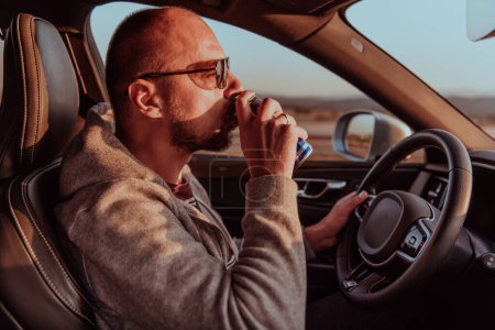 Téléchargez les photos : Un homme fatigué buvant un acoffee en conduisant une voiture au coucher du soleil. Voyage fatigué et long trajet. - en image libre de droit