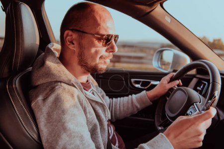 Photo for A man with a sunglasses driving a car and type a message on smartphone at sunset. The concept of car travel. - Royalty Free Image