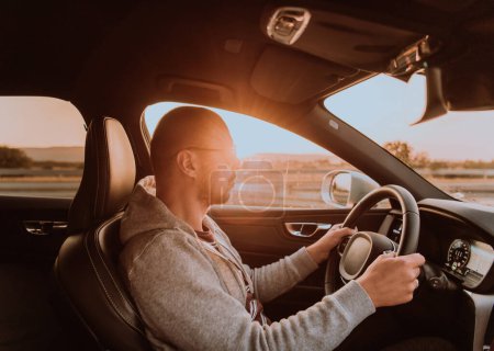 Téléchargez les photos : Un homme avec des lunettes de soleil conduisant une voiture au coucher du soleil. Le concept de voyage en voiture. - en image libre de droit