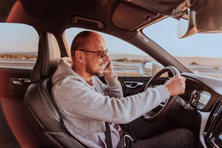 Photo for A man with a sunglasses driving a car and talking on smartphone at sunset. The concept of car travel. - Royalty Free Image