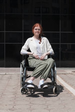 Foto de Frente a un moderno edificio corporativo, una joven sentada en una silla de ruedas con confianza, que simboliza el empoderamiento, la inclusión y la fuerza para superar los desafíos en el mundo de los negocios. - Imagen libre de derechos
