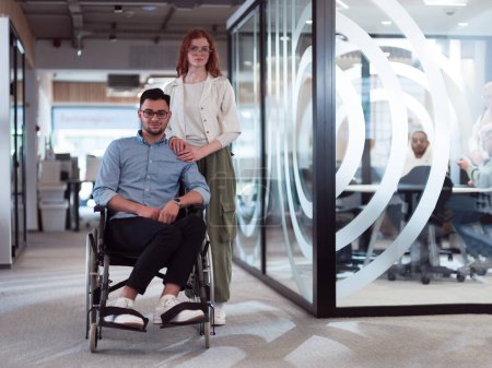 Foto de Jóvenes colegas de negocios, colegas de negocios colaborativos, incluida una persona en silla de ruedas, pasan por un moderno pasillo de oficinas de vidrio, ilustrando la diversidad, el trabajo en equipo y el empoderamiento en el - Imagen libre de derechos