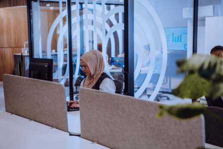Foto de En una oficina moderna, una joven empresaria musulmana que usa un hiyab se sienta con confianza y trabaja diligentemente en su computadora, encarnando determinación, creatividad y empoderamiento en el mundo de los negocios. - Imagen libre de derechos