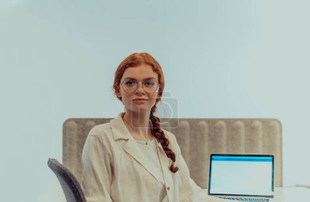 Téléchargez les photos : Une femme d'affaires moderne aux cheveux orange frappants assise en toute confiance dans un bureau contemporain, accompagnée de son ordinateur portable, incarnant le professionnalisme, la créativité et le succès dans une technologie axée sur - en image libre de droit