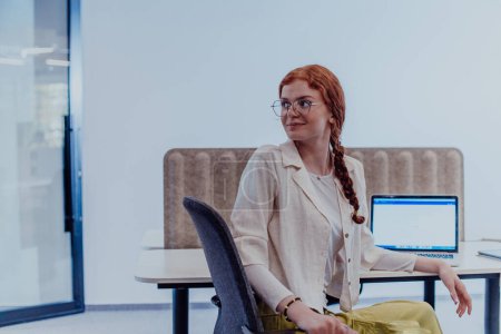 Téléchargez les photos : Une femme d'affaires moderne aux cheveux orange frappants assise en toute confiance dans un bureau contemporain, accompagnée de son ordinateur portable, incarnant le professionnalisme, la créativité et le succès dans une technologie axée sur - en image libre de droit