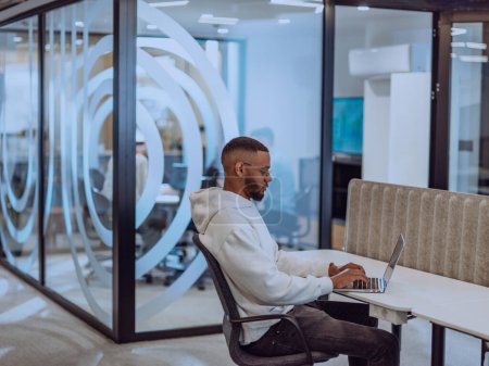 Téléchargez les photos : Dans un environnement de bureau moderne, un homme d'affaires afro-américain travaille diligemment sur son ordinateur portable, incarnant la détermination, l'ambition et la productivité dans son environnement professionnel.. - en image libre de droit