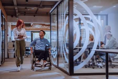 Foto de Jóvenes colegas de negocios, colegas de negocios colaborativos, incluida una persona en silla de ruedas, pasan por un moderno pasillo de oficinas de vidrio, ilustrando la diversidad, el trabajo en equipo y el empoderamiento en el - Imagen libre de derechos