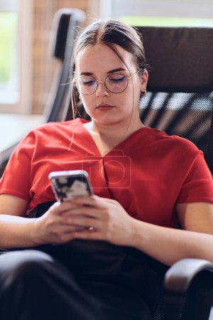 Téléchargez les photos : Une femme d'affaires se reposant sur une courte pause du travail dans un centre de coworking de démarrage moderne, en utilisant son smartphone pour se détendre et se recharger - en image libre de droit