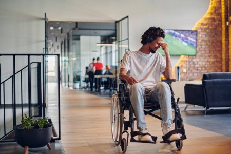 Foto de Un triste hombre de negocios en silla de ruedas ocupa un pasillo dentro de un moderno centro de coworking de startups, encarnando inclusividad y determinación en el entorno empresarial. - Imagen libre de derechos