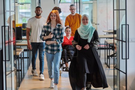 Photo for A diverse group of business people walking a corridor in the glass-enclosed office of a modern startup, including a person in a wheelchair and a woman wearing a hijab, showing a dynamic mix of - Royalty Free Image