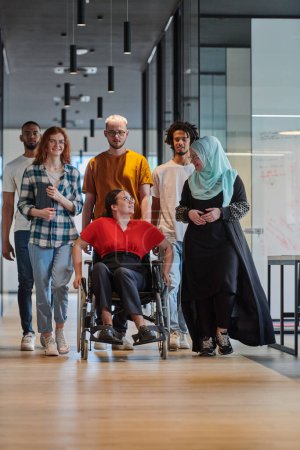 Téléchargez les photos : Un groupe diversifié de jeunes gens d'affaires marchant dans un couloir dans le bureau vitré d'une start-up moderne, y compris une personne en fauteuil roulant et une femme portant un hijab, montrant un mélange dynamique de - en image libre de droit