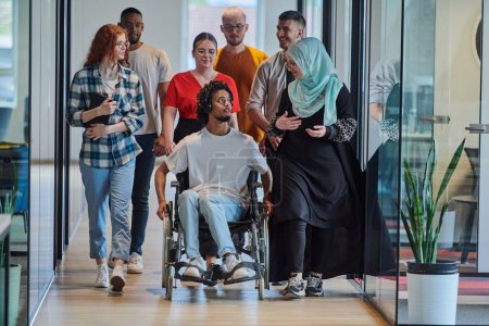 Un groupe diversifié de jeunes gens d'affaires marchant dans un couloir dans le bureau vitré d'une start-up moderne, y compris une personne en fauteuil roulant et une femme portant un hijab, montrant un mélange dynamique de