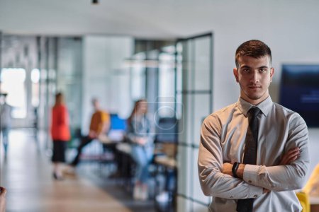 Téléchargez les photos : Un jeune chef d'entreprise se tient les bras croisés dans un couloir de bureau moderne, rayonnant de confiance et d'un sens de l'objectif, incarnant une présence dynamique et inspirante - en image libre de droit