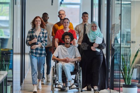 Téléchargez les photos : Un groupe diversifié de jeunes gens d'affaires marchant dans un couloir dans le bureau vitré d'une start-up moderne, y compris une personne en fauteuil roulant et une femme portant un hijab, montrant un mélange dynamique de - en image libre de droit