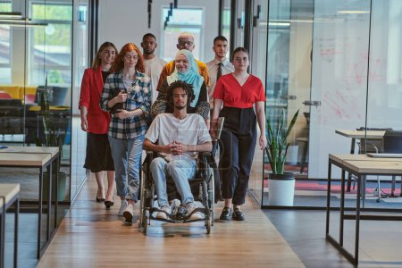 Foto de Un grupo diverso de jóvenes empresarios que caminan por un pasillo en la oficina acristalada de una startup moderna, incluyendo una persona en silla de ruedas y una mujer que lleva un hijab, mostrando una mezcla dinámica de - Imagen libre de derechos