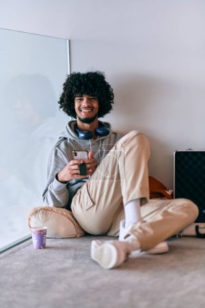 Téléchargez les photos : Entrepreneur afro-américain prenant une pause détente du travail, assis sur le sol tout en utilisant un casque sans fil et un smartphone pour certains divertissements numériques - en image libre de droit
