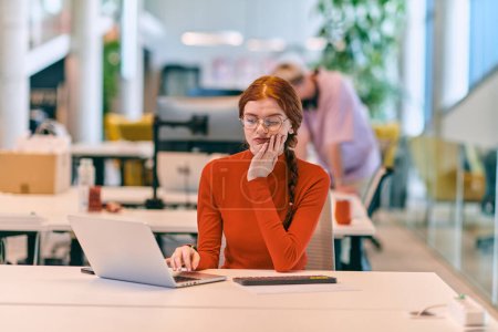 Téléchargez les photos : Dans un bureau de start-up moderne, une femme d'affaires professionnelle aux cheveux oranges assise à son ordinateur portable, incarnant l'innovation et la productivité dans son espace de travail contemporain - en image libre de droit