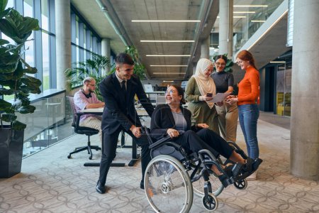 Foto de Un grupo diverso de colegas de negocios se está divirtiendo con su colega que usa sillas de ruedas, demostrando su atención e inclusión en el lugar de trabajo. - Imagen libre de derechos