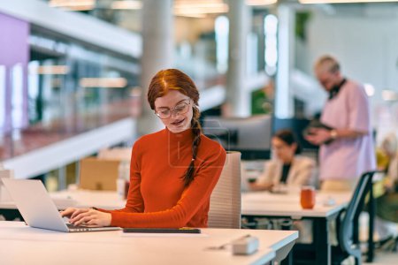 Foto de En una moderna startup, una empresaria profesional con el pelo naranja sentada en su portátil, personificando la innovación y la productividad en su espacio de trabajo contemporáneo - Imagen libre de derechos