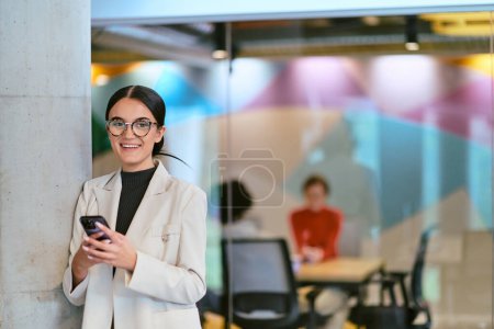 Foto de En una moderna oficina de vidrio, una mujer de negocios contemporánea sin esfuerzo multitareas, utilizando su teléfono inteligente, mientras está rodeada de colegas, que ilustran la dinámica de la vida corporativa y la tecnología - Imagen libre de derechos