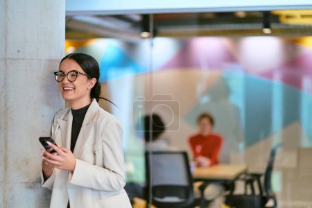 Foto de En una moderna oficina de vidrio, una mujer de negocios contemporánea sin esfuerzo multitareas, utilizando su teléfono inteligente, mientras está rodeada de colegas, que ilustran la dinámica de la vida corporativa y la tecnología - Imagen libre de derechos