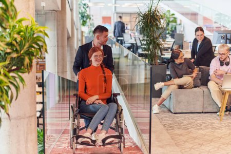 Téléchargez les photos : Un directeur d'entreprise aide son collègue d'affaires dans un fauteuil roulant, l'aidant à naviguer vers leur bureau de démarrage, où ils travaillent aux côtés de leur équipe diversifiée de collègues, mettant l'accent sur l'inclusivité et - en image libre de droit
