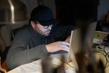 Photo for A close-up shot of a man using a laptop in a dimly lit room, engrossed in his digital work during the late hours of the night. - Royalty Free Image