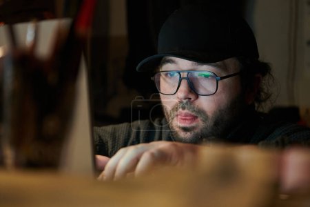 Foto de Un primer plano de un hombre usando un portátil en una habitación débilmente iluminada, absorto en su trabajo digital durante las últimas horas de la noche. - Imagen libre de derechos