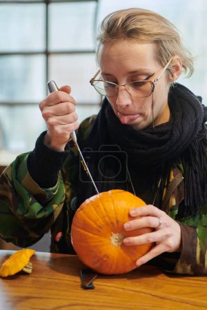 Téléchargez les photos : Une femme blonde moderne en uniforme militaire sculpte des citrouilles effrayantes avec un couteau pour la nuit d'Halloween. - en image libre de droit