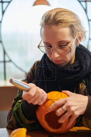Téléchargez les photos : Une femme blonde moderne en uniforme militaire sculpte des citrouilles effrayantes avec un couteau pour la nuit d'Halloween. - en image libre de droit