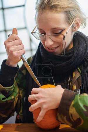 Foto de Una mujer rubia moderna en uniforme militar está tallando calabazas espeluznantes con un cuchillo para la noche de Halloween. - Imagen libre de derechos