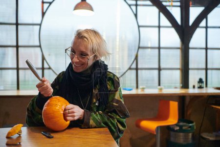 Photo for A modern blonde woman in military uniform is carving spooky pumpkins with a knife for Halloween night. - Royalty Free Image