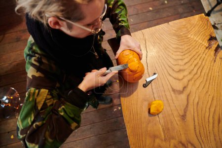 Foto de Una mujer rubia moderna en uniforme militar está tallando calabazas espeluznantes con un cuchillo para la noche de Halloween. - Imagen libre de derechos