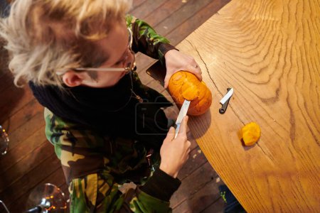 Photo for A modern blonde woman in military uniform is carving spooky pumpkins with a knife for Halloween night. - Royalty Free Image