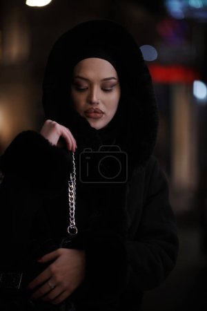 Foto de Mujer musulmana caminando en una calle urbana en una fría noche de invierno usando hijab con luces bokeh de la ciudad en el fondo - Imagen libre de derechos